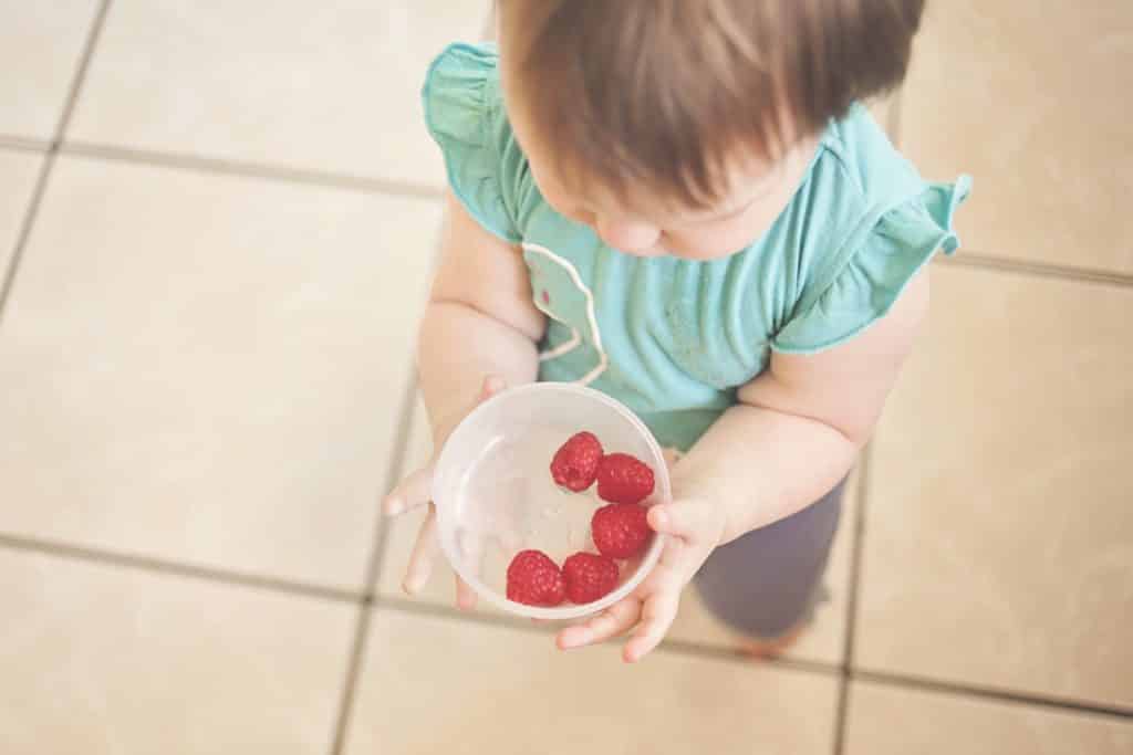 White appearing child in blue frilled t-shirt and jeans holding a bowl of raspberries