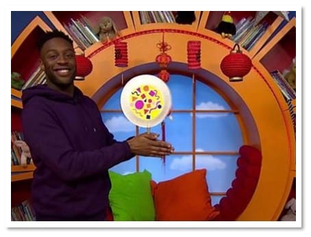 Black TV presenter with short hair wearing a purple hoodie and smiling by an orange window, holding a Chinese rattle drum.