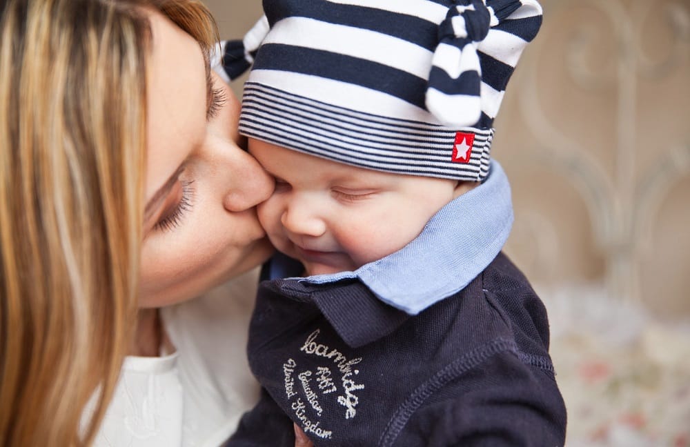 White mother kissing her baby who is wearing a blue and white stripey hat and blue coat