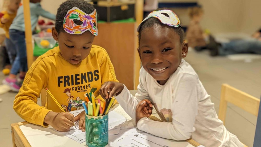 two young black children taking part in a mask making activity. Young girls is smiling at the camera