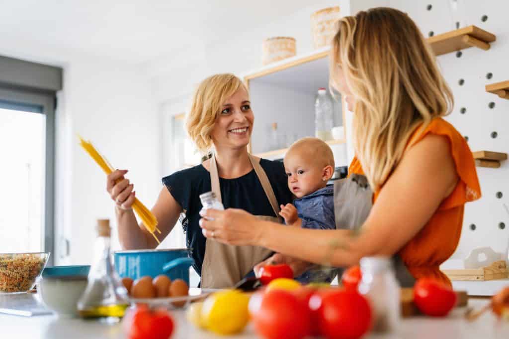 White female couple one holding their baby cooking with spaghetti and fresh vegetables