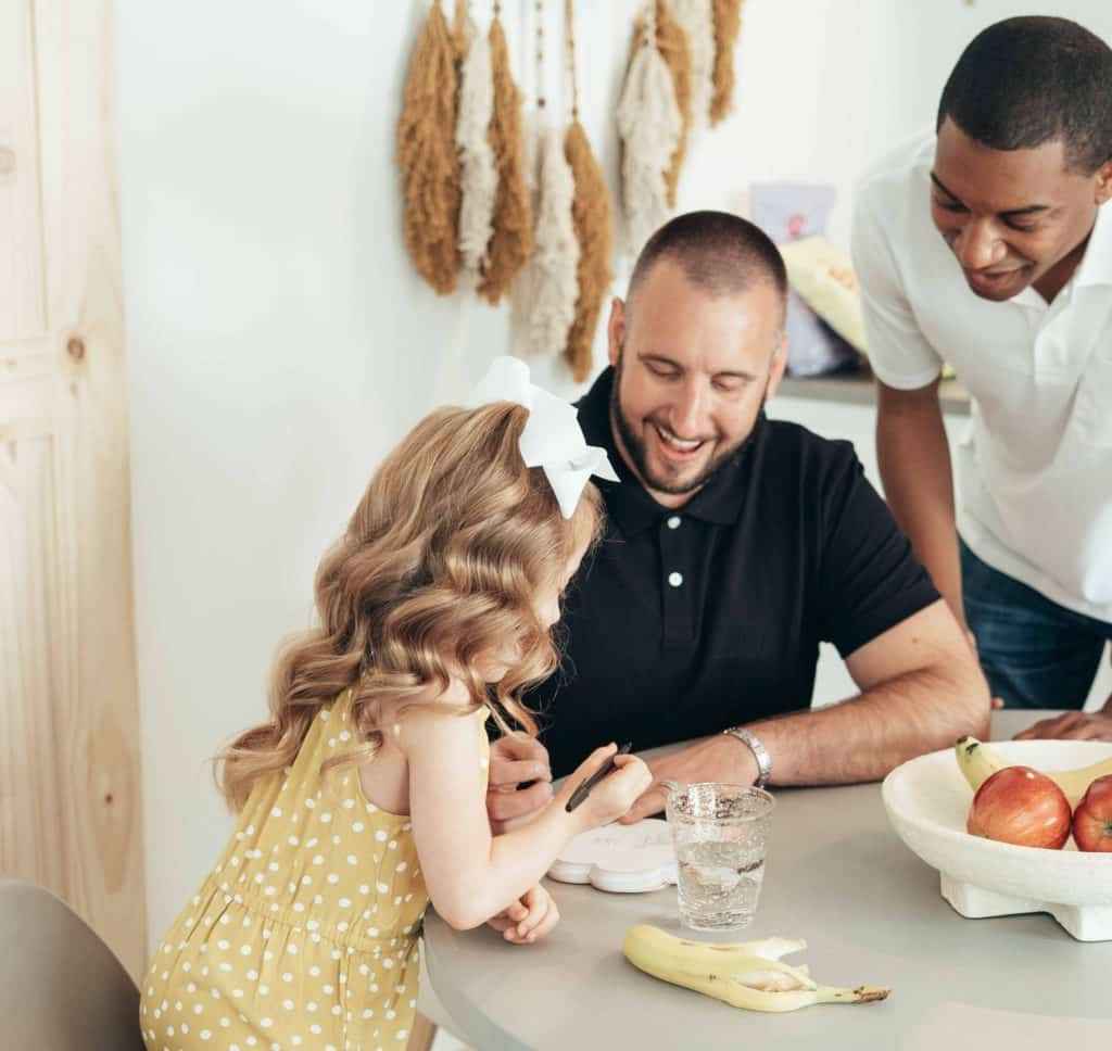 Black and white Dads in a white contemporary room drawing and smiling with their daughter 