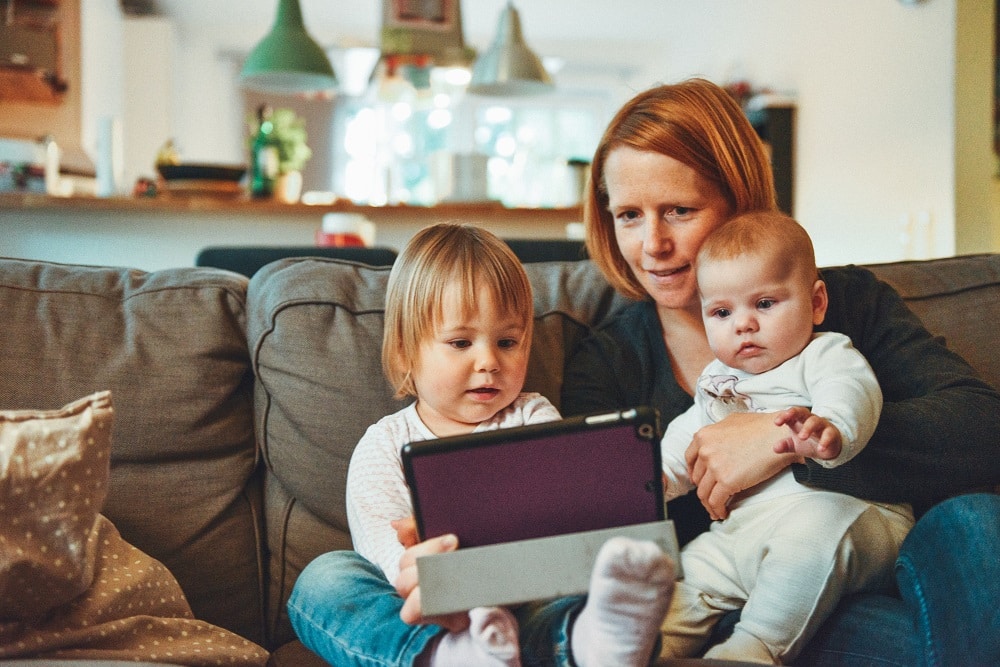 White appearing ginger haired young mother sitting on sofa with baby on her lap looking at table with toddler