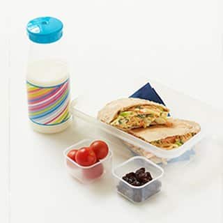 Photo of a lunch box containing a cheese and coleslaw pitta, a tub containing cherry tomatoes, a tub containing raisins and a small drink bottle filled with milk