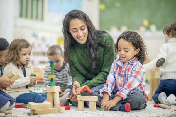 A boy and his teacher are indoors in their kindergarten classroom. They are building with blocks together.