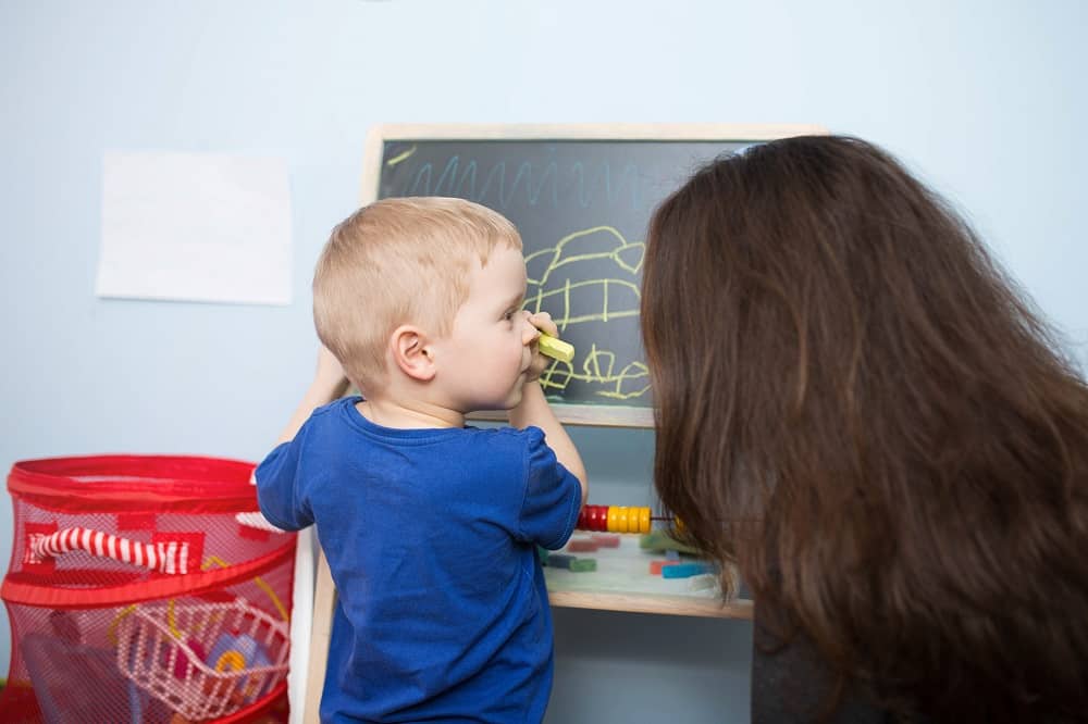 A photo of small boy drawing on the blackboard and his big sister looking after him. They're in children's room.