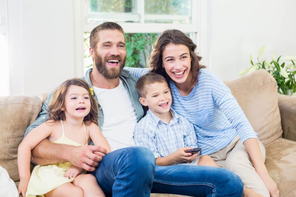 White family, smiling casually dressed sitting on a sofa. Mum, Dad, son and daughter