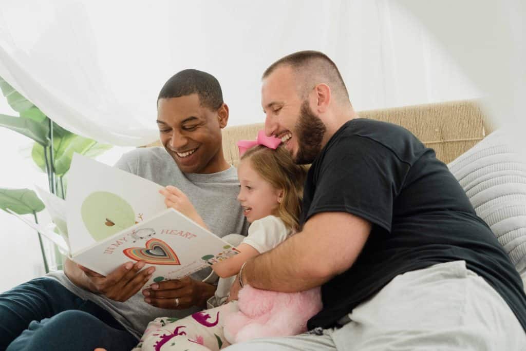 Two gay dads one black and one white with their white daughter smiling and reading a book.