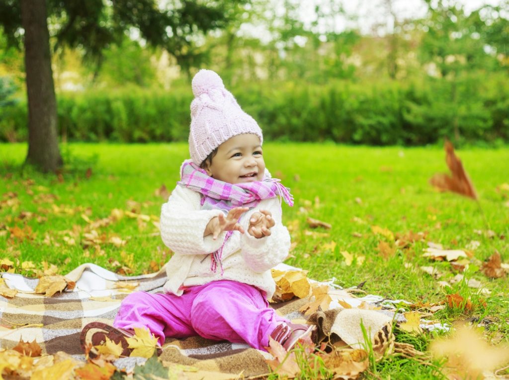 happy little girl in the autumn park playing with leaves in pink bobble hat, scarf, trousers and trousers