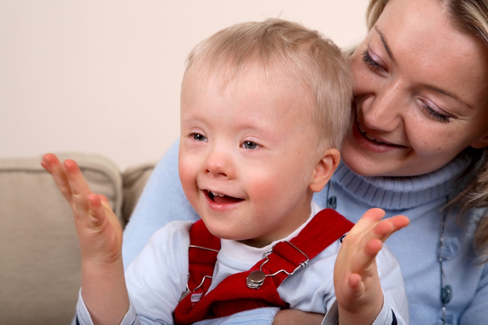 A close-up portrait of a white mother smiling holding her white smiling and happy young son in red dungarees with Down Syndrome.