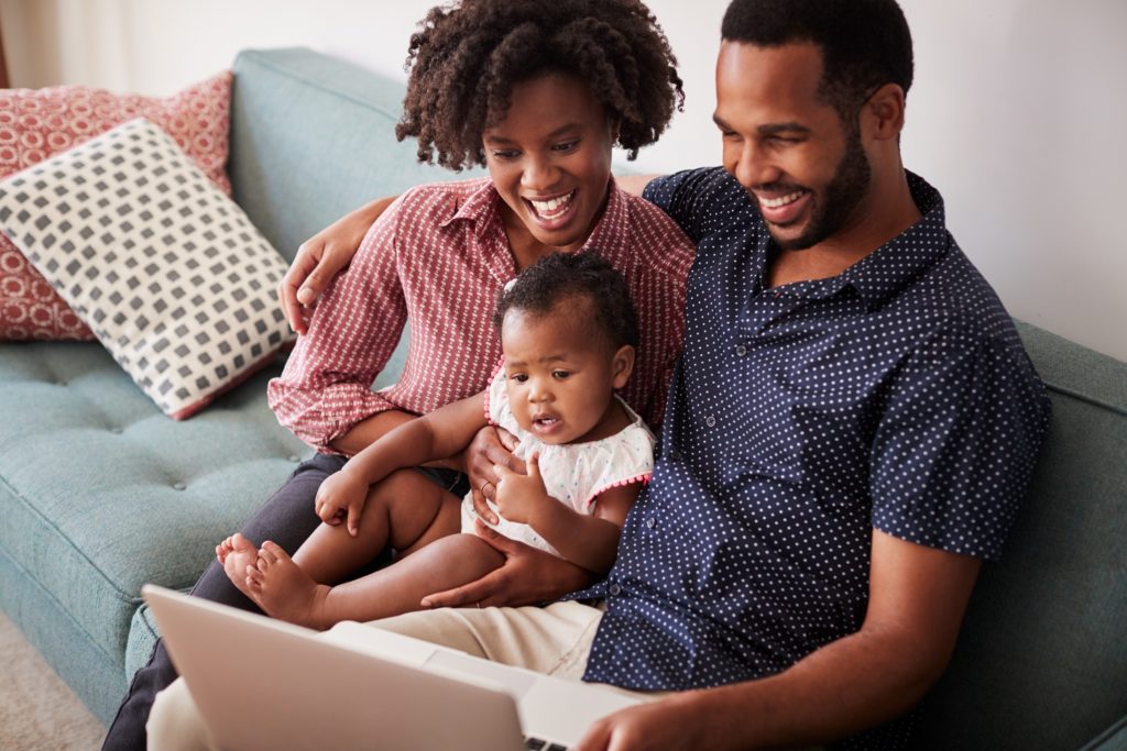 Black Family of male and female partners With Baby Daughter Sitting On Sofa At Home Looking At Laptop Computer