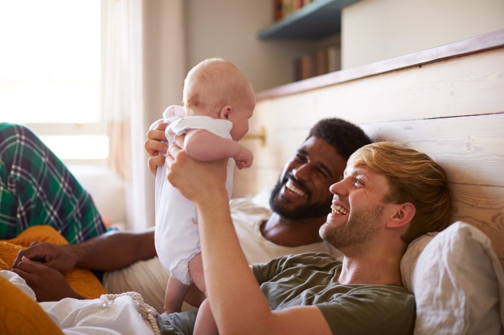 Two happy Gay dads smiling - one black and one white laying fully dressed on a bed and holding up their baby 