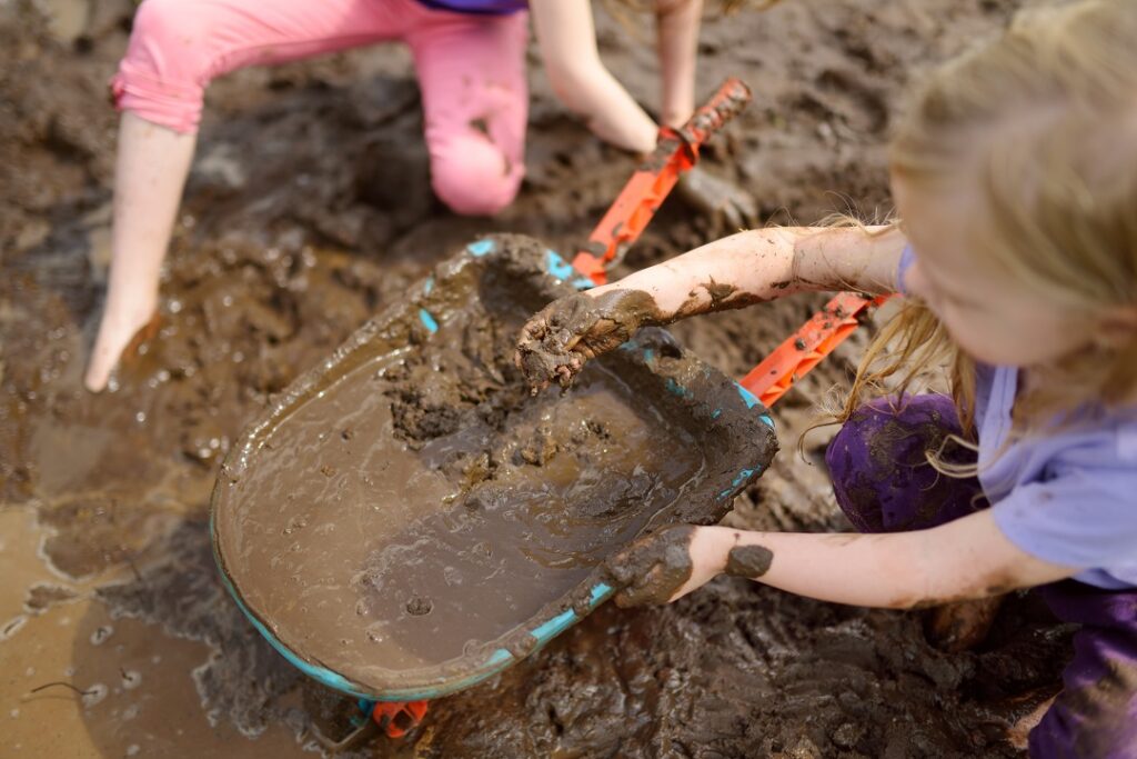 Two white toddlers playing with mud in a blue and orange wheelbarrow