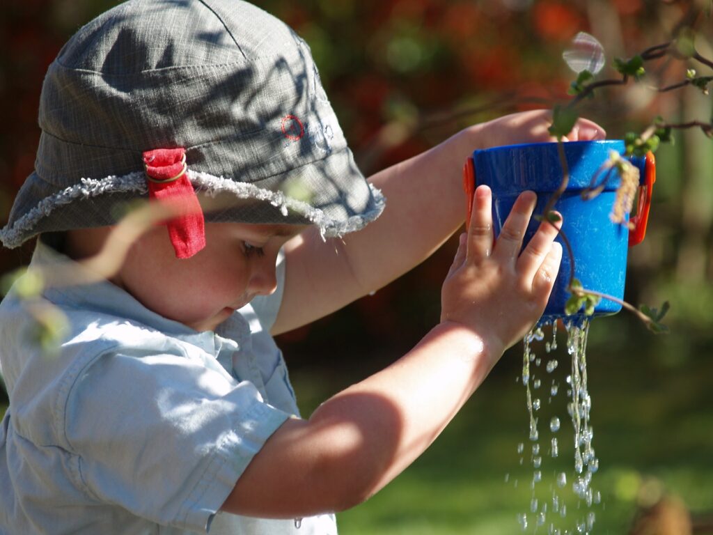 White toddler boy in grey havt watching water drip through a plant in a blue pot