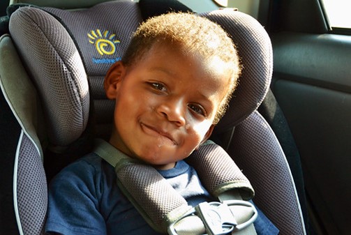 boy in blue hat in car seat in the car smiling at the camera