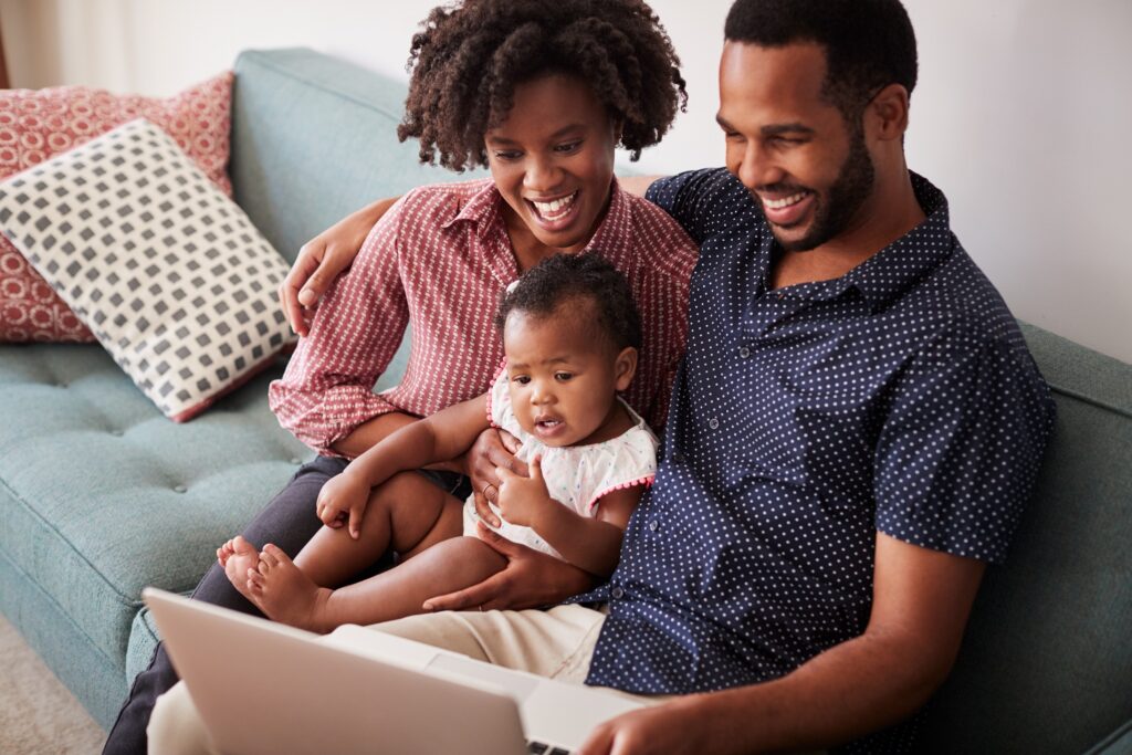 Black family of Mum, Dad With Baby Daughter Sitting On Sofa At Home Looking At Laptop Computer